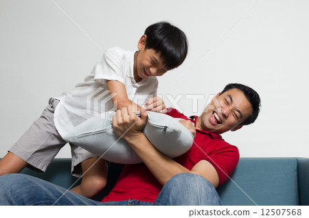 Asian boy is playing - pillow fight - with his dad / father on the sofa in the living room 12507568