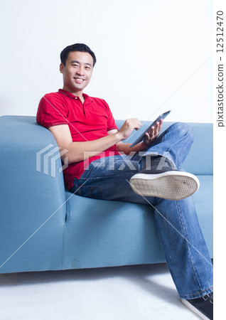 A young Asian man enjoy the moment using his mobile device while seating in his living room 12512470