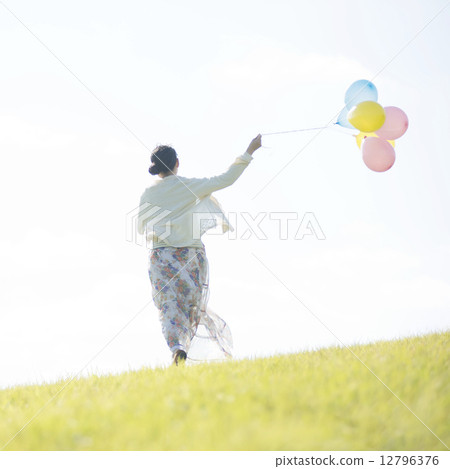 A rear view of a woman holding a balloon and running on the meadow 12796376