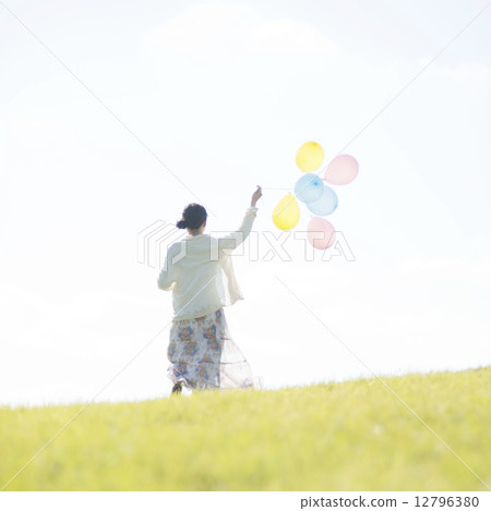 A rear view of a woman holding a balloon and running on the meadow 12796380