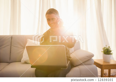 Happy man sitting on couch phoning and using laptop Happy man sitting on couch phoning and using laptop 12839101