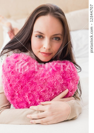Pretty brunette holding heart cushion on bed 12844707