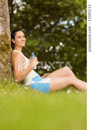 Cheerful fit brunette sitting against a tree holding a bottle 12985211