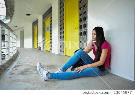 A sad and lonely young Asian female student seating by herself at a corridor of a campus A sad and lonely young Asian female student seating by herself at a corridor of a campus 13045977