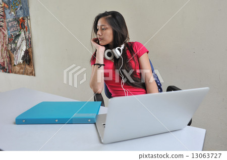 A young and beautiful Asian female student pondering / brainstorming at her desk in front of laptop A young and beautiful Asian female student pondering / brainstorming at her desk in front of laptop 13063727