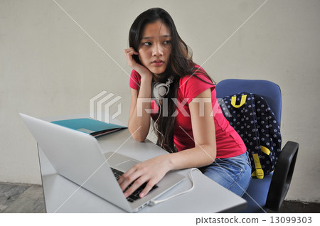 A young beautiful Asian female student looking restless seated at her desk in front of laptop 13099303