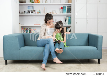 Young chinese mother and daughter using mobile device tablet iPad relaxing on a sofa 13218851