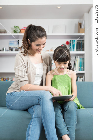 Young chinese mother and daughter using mobile device tablet iPad relaxing on a sofa 13218875