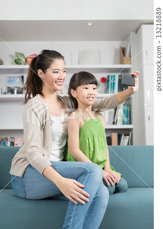 Young chinese mother and daughter taking selfies using mobile device smart phone relaxing on a sofa 13218889