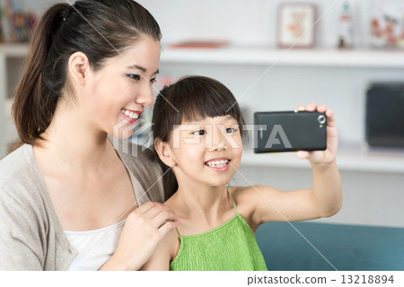 Young chinese mother and daughter taking selfies using mobile device smart phone in the living room 13218894