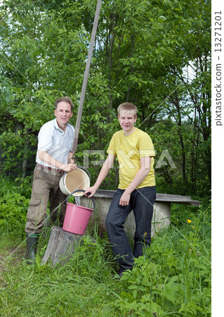 The boy helps the father to pour water in a bucket from a well.. The boy helps the father to pour water in a bucket from a well.. 13271201