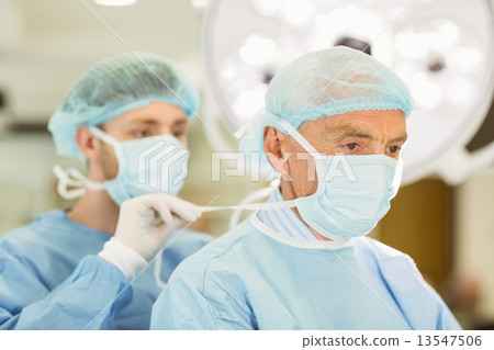 Stock Photo: Young surgeon tying older surgeons mask