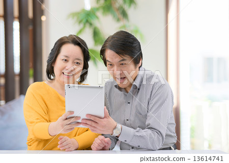 A middle-aged couple in casual wear, which is manipulating the tablet in the living room of the home A middle-aged couple in casual wear, which is manipulating the tablet in the living room of the home 13614741
