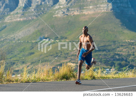 Athletic, muscular, healthy black male running along a road outdoors with a mountain background. 13626683