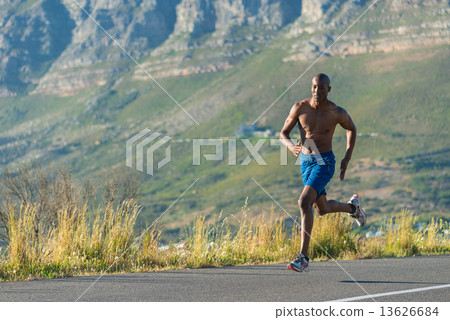 Athletic, muscular, healthy black male running along a road outdoors with a mountain background. 13626684