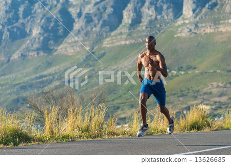 Athletic, muscular, healthy black male running along a road outdoors with a mountain background. 13626685