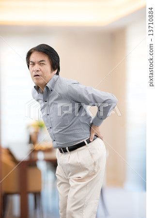 Middle-aged man in casual wear suffering from back pain at home living Middle-aged man in casual wear suffering from back pain at home living 13710884