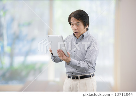 A middle-aged man with casual wear manipulating the tablet at the window of the home living room 13711044