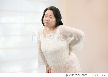 A middle-aged woman in casual wear suffering from low back pain, stiff shoulder standing in the room 13711127