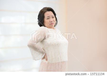 A middle-aged woman in casual wear suffering from low back pain, stiff shoulder standing in the room 13711134