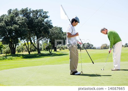 Golfer holding hole flag for friend putting ball Golfer holding hole flag for friend putting ball 14087665