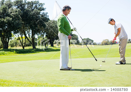 Golfer holding hole flag for friend putting ball Golfer holding hole flag for friend putting ball 14088780