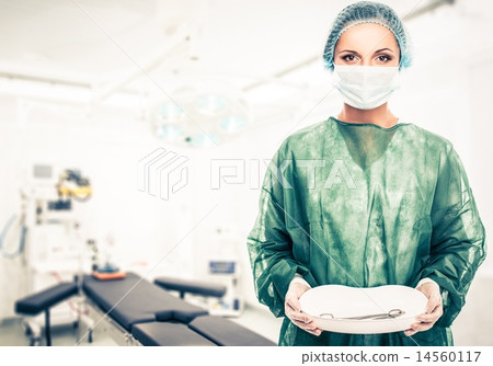 Young woman doctor in cap and face mask holding medicine tray with surgery tools Young woman doctor in cap and face mask holding medicine tray with surgery tools 14560117