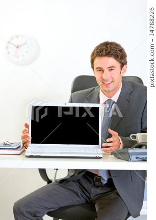 Smiling businessman sitting at office desk and showing laptops b 14788226