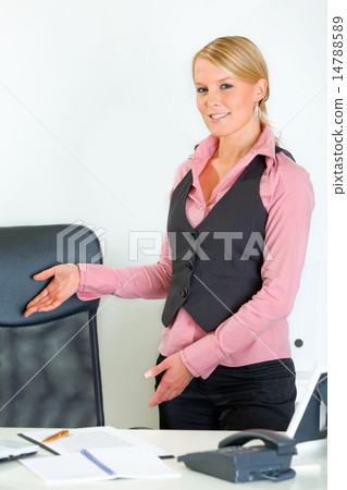 Smiling business woman standing near office desk and inviting to sit on chair. 14788589