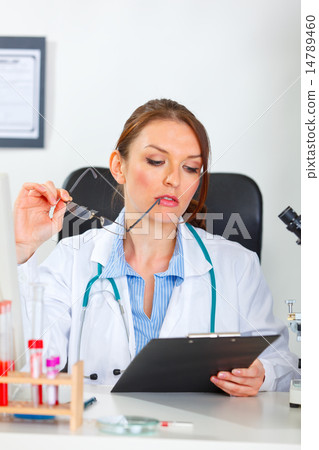 Thoughtful female medical doctor looking in clipboard at cabinet Thoughtful female medical doctor looking in clipboard at cabinet 14789460