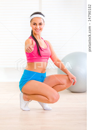 Smiling fit young girl squatting down and showing thumbs up gesture. 14790182