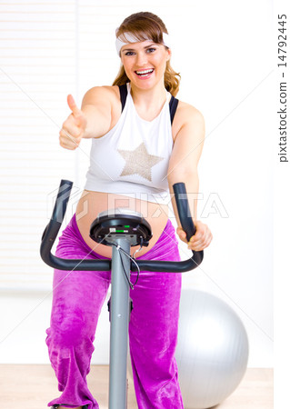Smiling beautiful pregnant woman working out on static bicycle and showing thumbs up gesture. 14792445