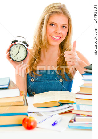 Smiling teengirl with alarm clock sitting at table and showing thumbs up gesture. 14793233
