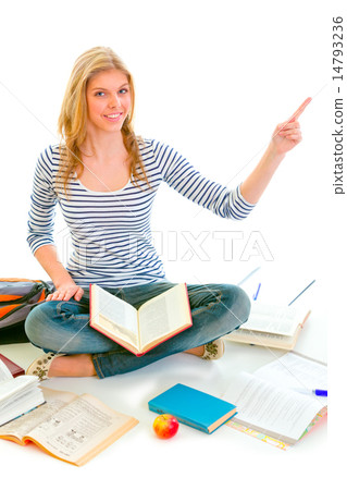 Smiling teen girl sitting on floor among schoolbooks and pointin 14793236