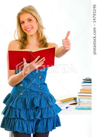 Smiling girl with book in hand standing near table and showing thumbs up gesture. 14793246