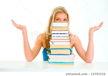 Sitting at table confused teen girl spreading hands apart and looking out of pile of book. 14793258