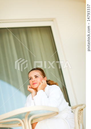 Dreaming young woman in bathrobe sitting at table on terrace 14793337