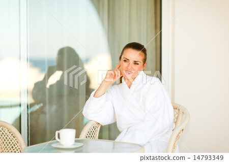 Thoughtful pretty woman in bathrobe sitting at table on terrace 14793349