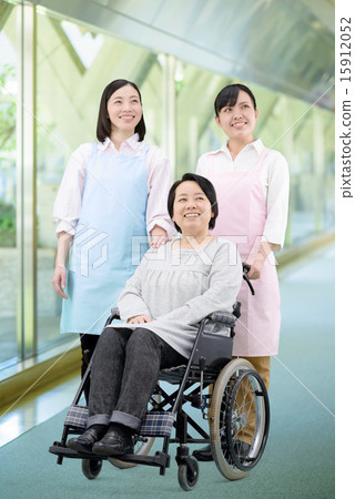 Two female helper in the apron in the corridor walking along the facility Image of the woman in the wheelchair Two female helper in the apron in the corridor walking along the facility Image of the woman in the wheelchair 15912052