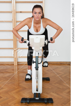 fit woman on stationary bicycle in gym 16489619