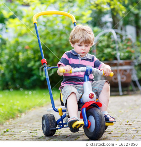 Active blond kid boy driving tricycle or bicycle in domestic gar 16527850