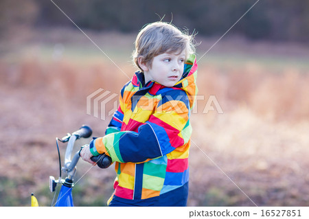 kid boy in safety helmet and colorful raincoat riding bike, outd 16527851