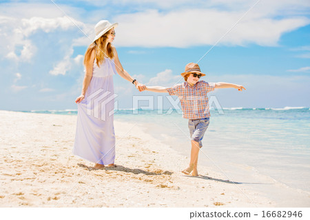 Happy beautiful mother in long white dress enjoying beach time with her son wearing hat and 16682946