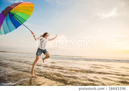 Cheerful young girl with rainbow umbrella having fun on the beach before sunset. Travel, holidays Cheerful young girl with rainbow umbrella having fun on the beach before sunset. Travel, holidays 16683396
