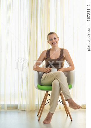 Happy young woman sitting on modern chair with cup of tea Happy young woman sitting on modern chair with cup of tea 16725141