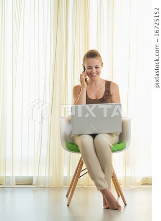 Smiling young woman on modern chair with laptop talking mobile p Smiling young woman on modern chair with laptop talking mobile p 16725152