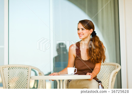 Happy young woman sitting on terrace with cup of tea and looking 16725628