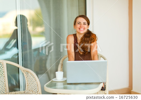 Happy young woman sitting on terrace with cup of coffee and lapt 16725630