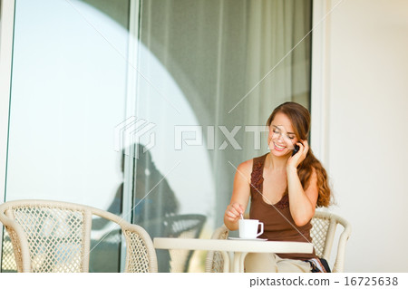 Happy young woman sitting on terrace with cup of coffee and spea 16725638