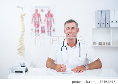 Smiling doctor writing on clipboard at his desk Smiling doctor writing on clipboard at his desk 17168857